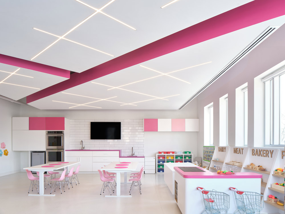 A classroom with two long kids' tables, the room is in predominantly white and pink colour scheme. Play stations on the right under the large windows, and a large TV on the wall above the kitchen area. Linear inset lighting on the ceiling above