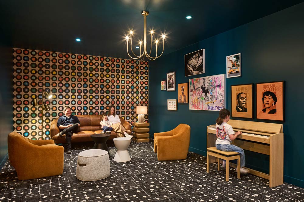 Music room designed in darker colour scheme with dark green walls, contrasting orange lounge chairs and sofa, and colourful art work. A girl is playing the piano while 2 adults are sitting on the sofa