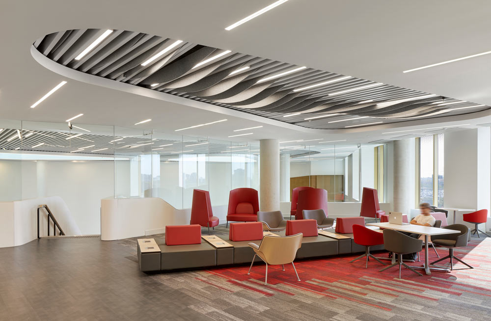 A lounge area adjacent to the atrium, with read lounge chairs and neutral flooring, and above is a slated detail on the ceiling above the lounge area