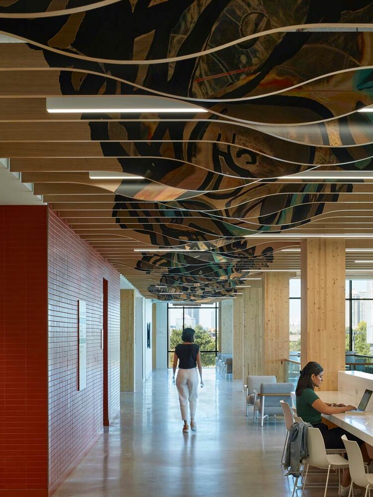 A hallway above which overlapping wavy wooden pieces with colourful indigenous art painted on it are hanging from the ceiling with linear lighting running in between them.