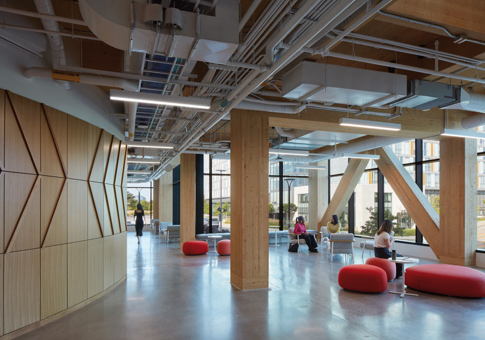Bright and open lounge area surrounded by large glass walls and views of the surrounding buildings and nature. The interior has a lot of wood elements and finishes, with exposed HVAC and pipes above 