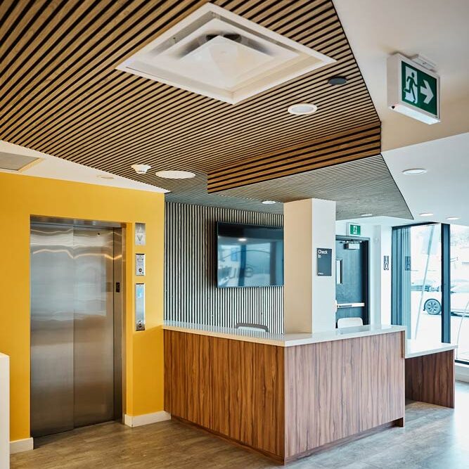 Front lobby with wooden slat detailing running along the wall behind the reception desk and up the ceiling. The elevator door is surrounded by a bright yellow wall