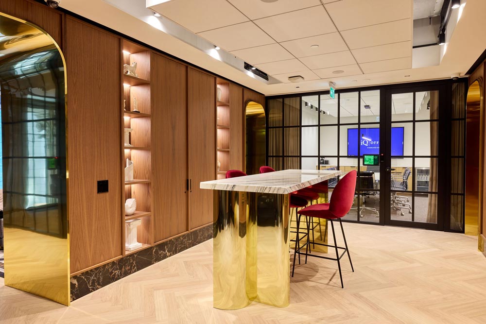 Another common area with a bar table and stools adjacent to another board room separated by glass walls with black metal frames. There are gold finishes throughout and wood panels on the walls