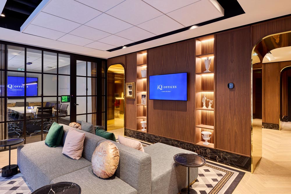 A common area with modern sofas in a space with wood panelled walls and lit built-ins. The space is connected to the next room by an archway dressed in gold material