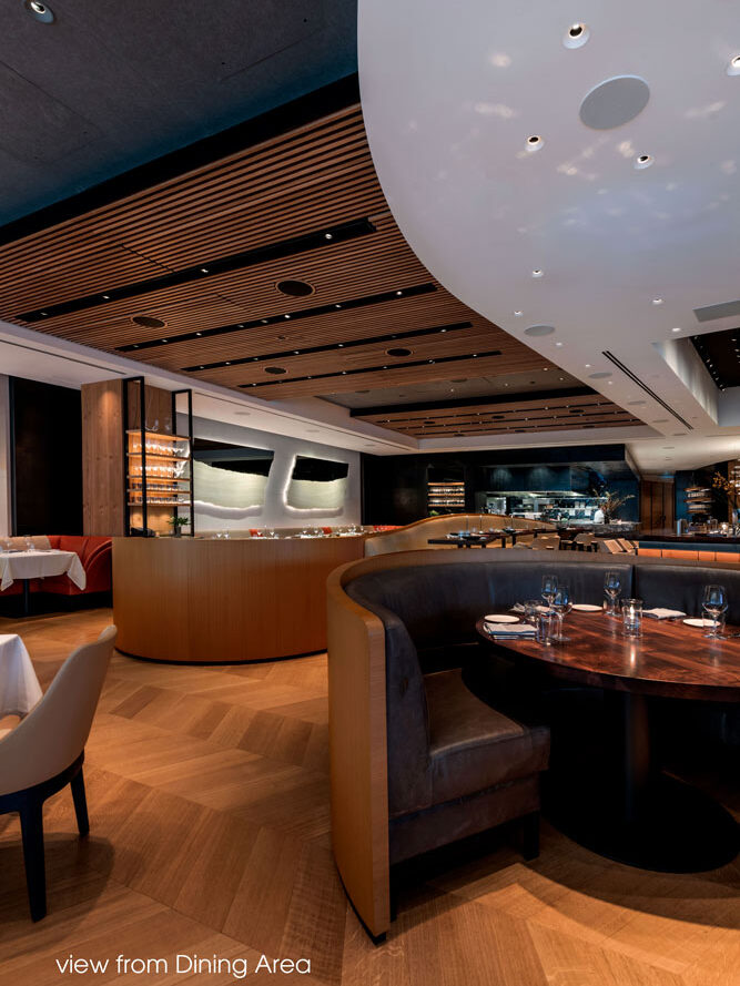 View from the dining area showing curved banquettes hugging round tables, and in the background is the sushi bar. The ceiling above is a combination of white rounded element with wood slatted detail