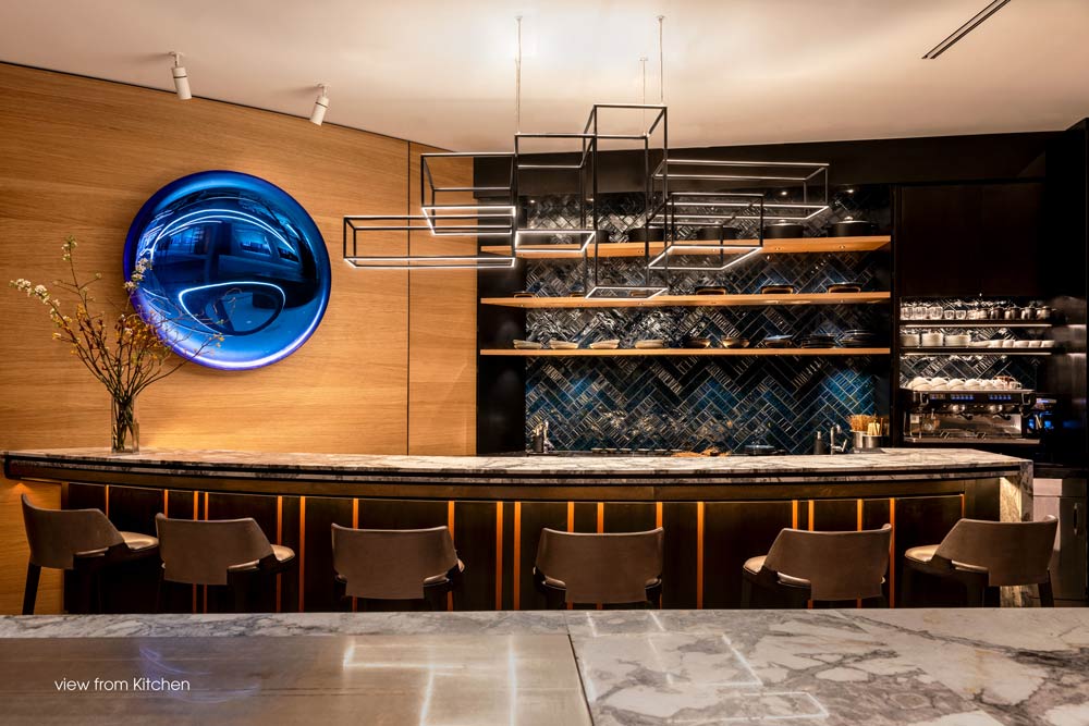 Sushi Bar with stools attached to the wood paneled wall that features a round blue decorative detail. A sculptural linear light fixture hangs above the bar