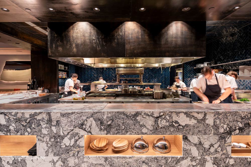 Open concept kitchen with a stone counter all around the perimeter of the kitchen, showing people cooking and a large industrial hood is in the centre