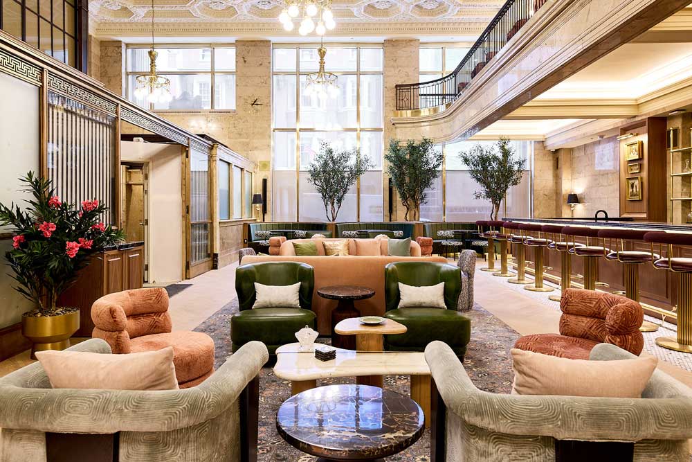 A view of the main lobby that looks more luxury hotel than an office, anchored by a curated barista bar, and in the central area are vintage inspired lounge and seating. At the other end of the space are large two level windows above which are original plaster details in the ceiling