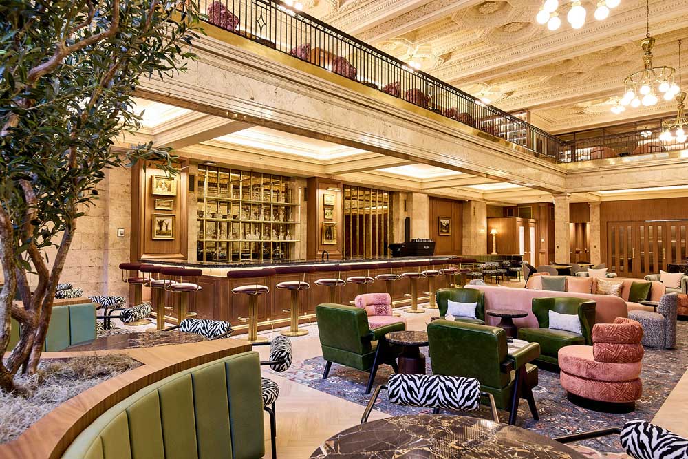 A view of the main lobby that looks more luxury hotel than an office, anchored by a curated barista bar, and in the central area are vintage inspired lounge and seating. The second level mezzanine is visible above the bar and also the decorative plaster ceilings