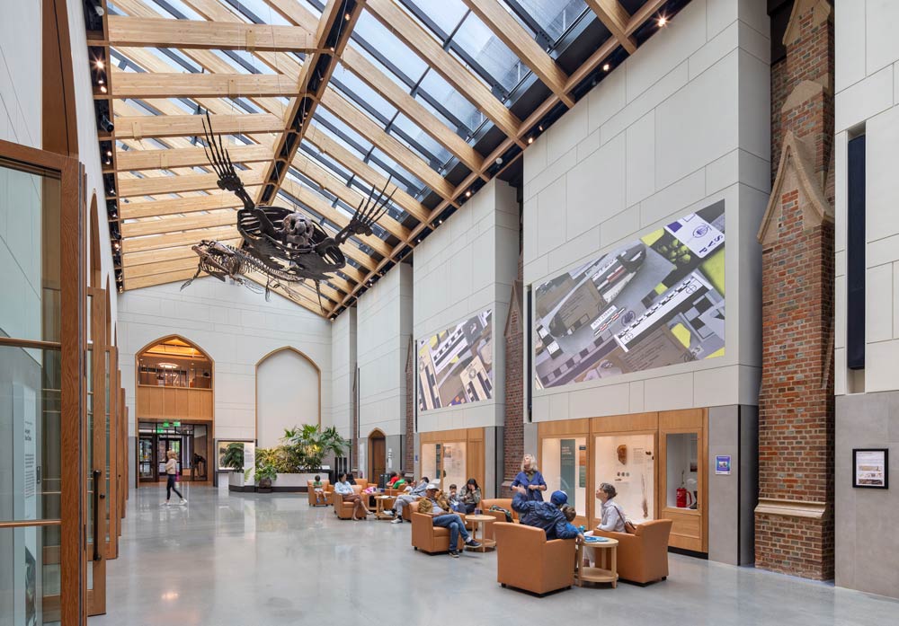 Central hall with lounge seating, it has tall ceiling with a skylight above underwhich a skeleton of a dinosaur hangs. Exposed brick is visible in the walls in contrast to modern, updated parts