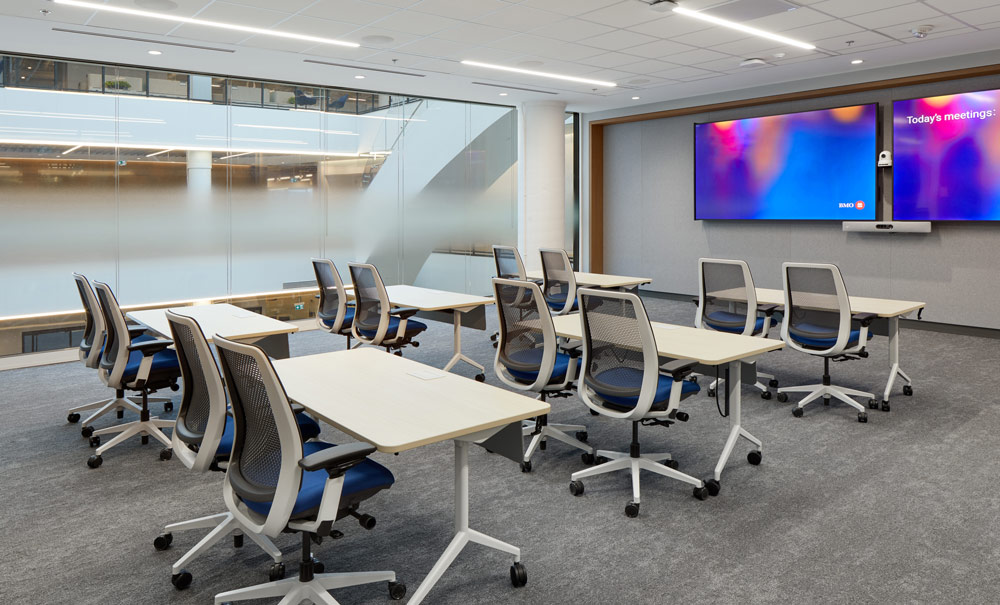 One of the lecture rooms behind a glass wall looking onto the atrium. Two large screens at the front wall and multiple desks for two