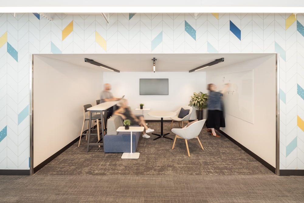 One of the areas meant for collaborative work sessions with sofa, lounge chairs and a bar height table in front of a large white board, and people sitting and working together