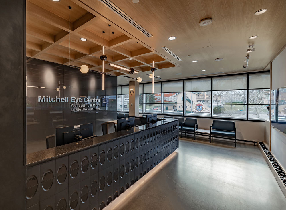 The front desk in the reception, where the reception desk is in black sleek finish, and so is the wall behind it where the name of the clinic is dispalyed. Above the reception desk area is a ceiling feature made of wooden squares from which light pendants are hanging