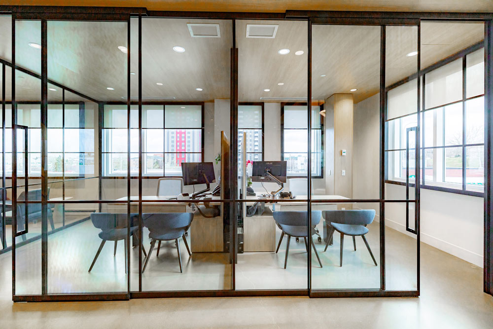 Two offices surrounded by windows are right next to each other, both behind glass walls with black metal framing