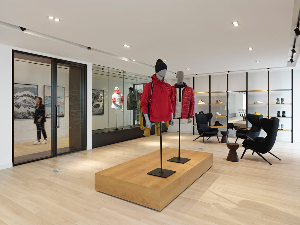A waiting area in a room where jackets and shoes are displayed on mannequins and on the wall shelfs, with a glass display window looking onto the hallway