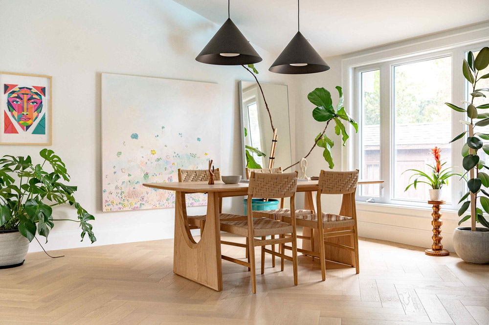 An open and bright dining room with a table in the centre with two black geometric pendants hanging above. Large art work hangs on the big wall behind the table, and largo windows on the right