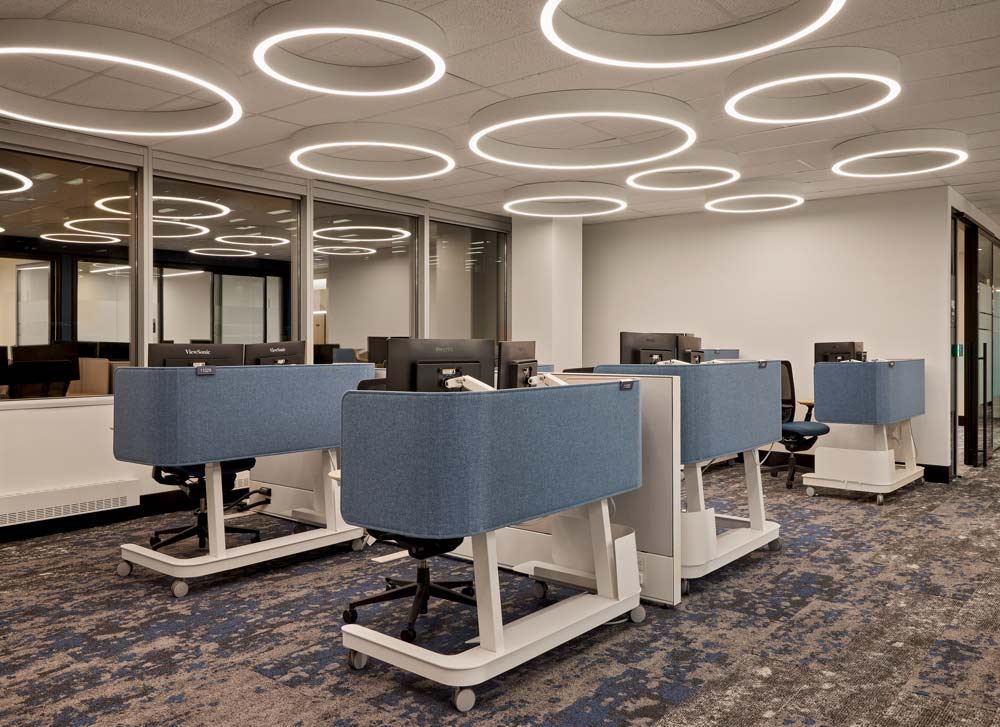 Work stations with light blue cubicle separators, and above are circular LED light fixtures