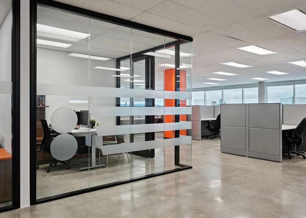 A private office is behind a glass wall, outside of which is the workstation area with cubicles