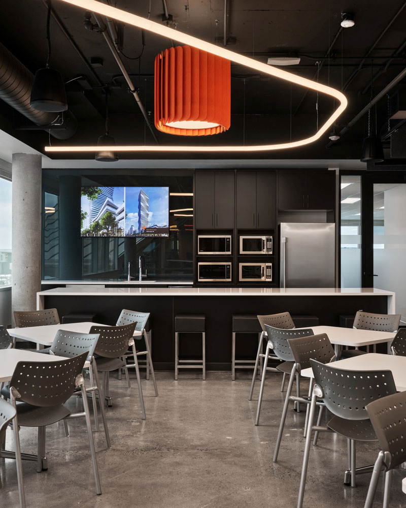 Kitchen done in black finishes, with a long island in front of it, and some tables for 4 . A large orange chandelier hangs above 