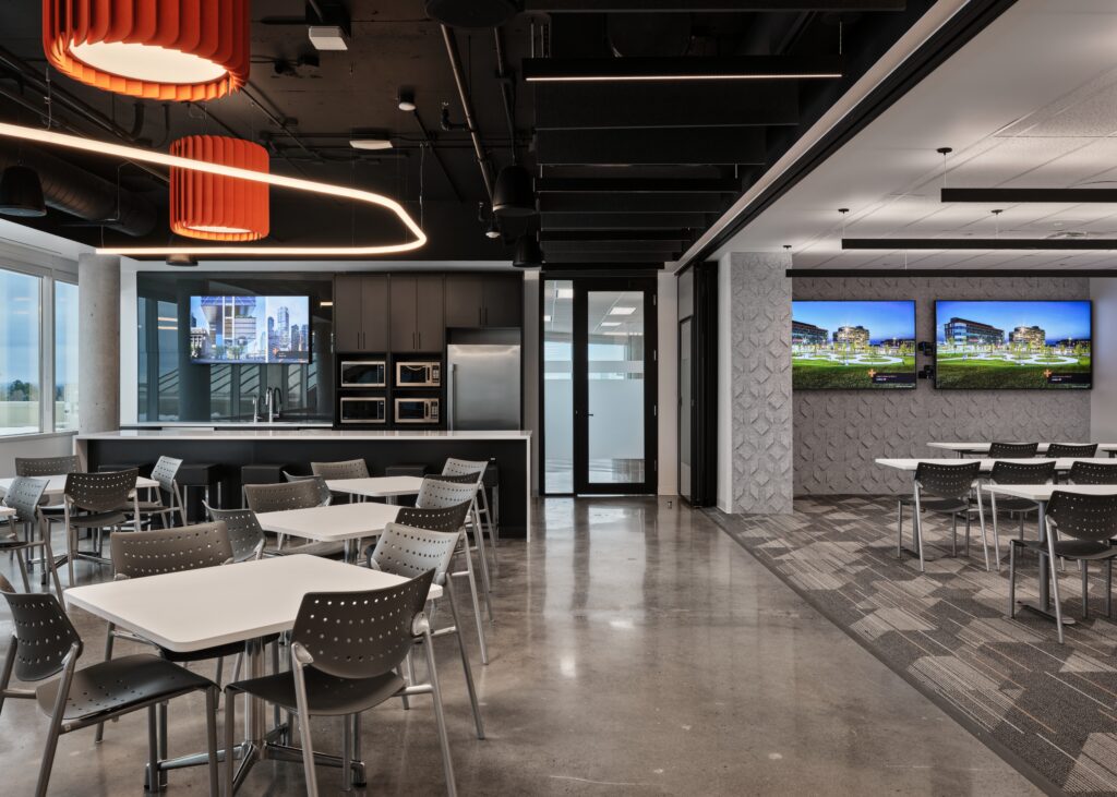 Open concept space with a cafeteria/kitchen area with exposed black ceilings and some orange details right next to the space with long tables in a few rows in front of 2 large TVs.