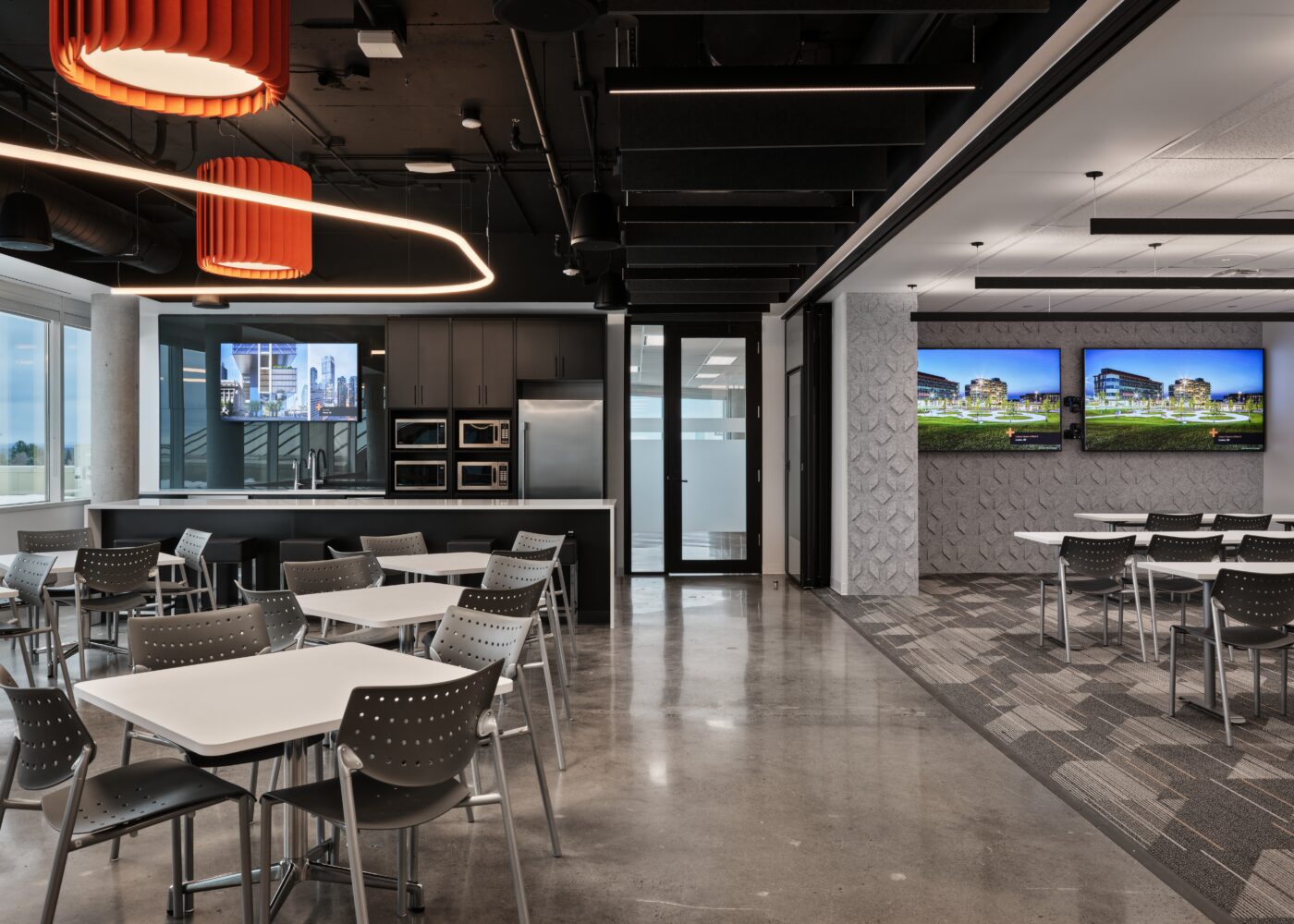 Open concept space with a cafeteria/kitchen area with exposed black ceilings and some orange details right next to the space with long tables in a few rows in front of 2 large TVs.