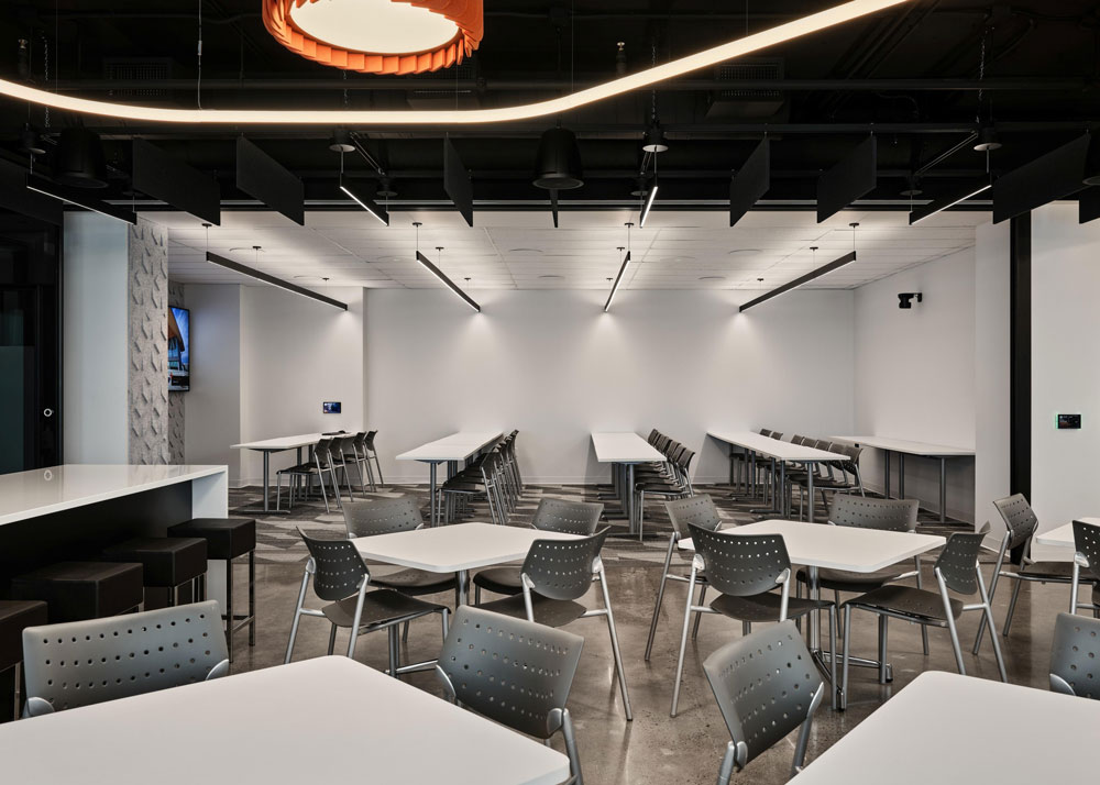 Cafeteria area with multiple desks in the front and in the back there are a few rows of long tables with chairs and linear minimalistic lighting above the tables